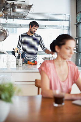 Buy stock photo Shot of a handsome young man preparing food in the kitchen while his wife is using a laptop