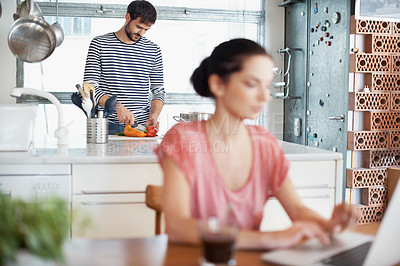 Buy stock photo Shot of a handsome young man preparing food in the kitchen while his wife is using a laptop