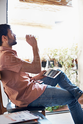Buy stock photo Shot of a handsome young man drinking a beer while sitting with his digital tablet indoors