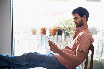 Buy stock photo Shot of a handsome young man relaxing with his digital tablet indoors
