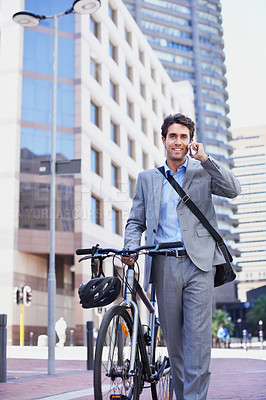 Buy stock photo A handsome businessman talking on his mobile while pushing his bicycle in the city central