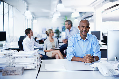 Buy stock photo Portrait of a smiling office worker sitting at his workstation with colleagues in the background