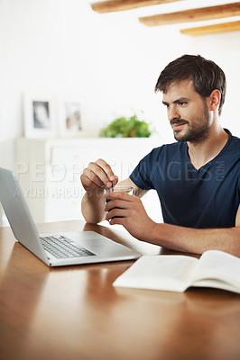 Buy stock photo A handsome young man working on his laptop while reading a book
