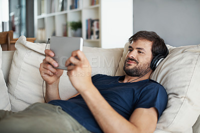 Buy stock photo A handsome young man listening to music on his tablet while lying on the sofa