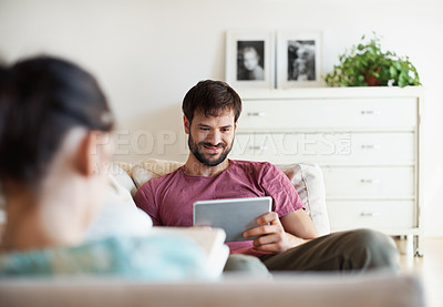 Buy stock photo A handsome young man using his tablet while his wife reads a book