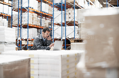 Buy stock photo A young man in a warehouse taking orders on his mobile phone and recording them on his laptop