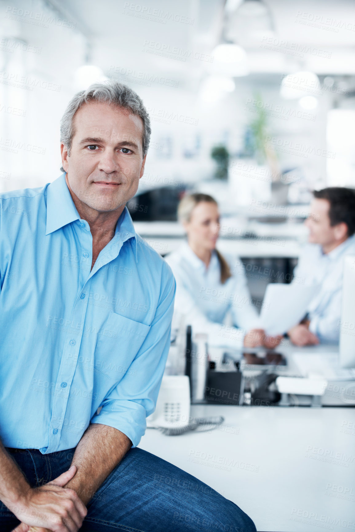 Buy stock photo Confident mature businessman sitting on a desk with his coworkers in the background