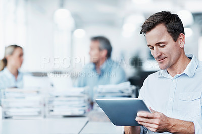 Buy stock photo A handsome young man using his digital tablet while two colleagues converse in the background