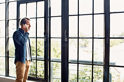 Buy stock photo Shot of a mature man talking on a cellphone while standing by windows in his apartment
