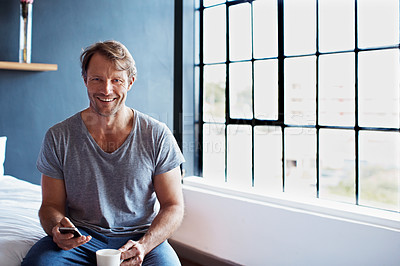 Buy stock photo Portrait of a smiling mature man sitting on his bed in the morning using a cellphone and drinking a cup of coffee