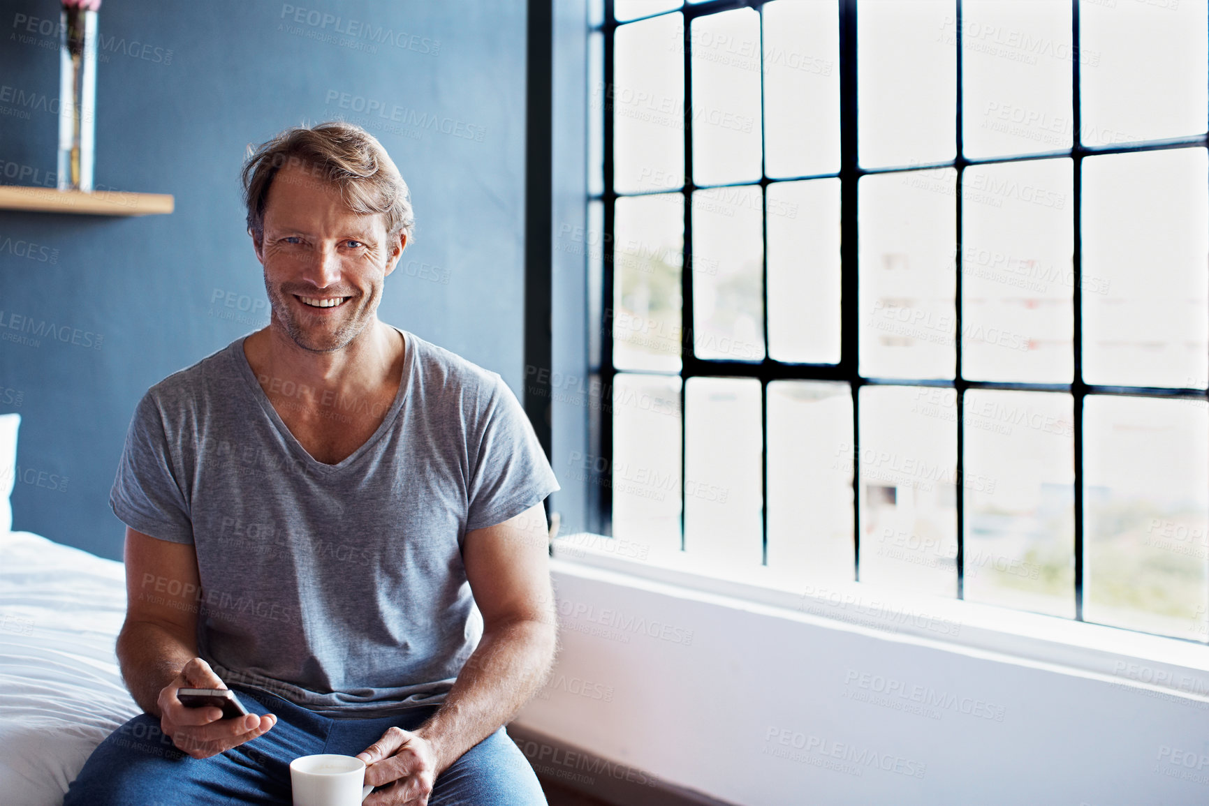 Buy stock photo Portrait of a smiling mature man sitting on his bed in the morning using a cellphone and drinking a cup of coffee