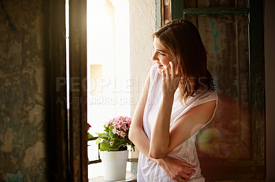 Buy stock photo Shot of an attractive young woman standing by a window at home talking on a cellphone