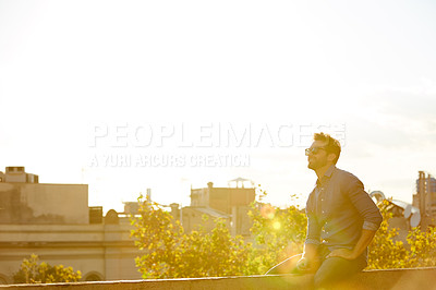 Buy stock photo Shot of a smiling young man sitting on a rooftop at sunset with the city in the background
