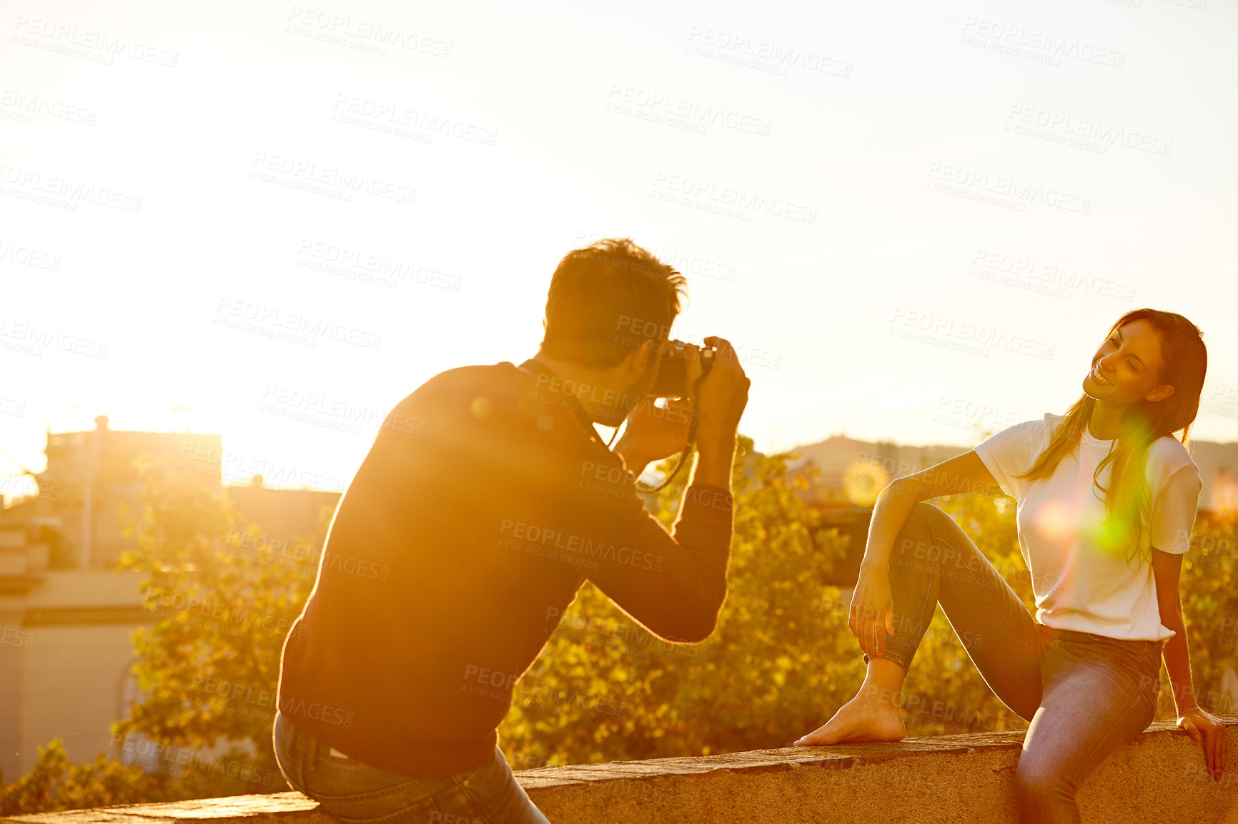 Buy stock photo Shot of a young man taking a photo of his girlfriend while sitting on a balcony overlooking the city at sunset
