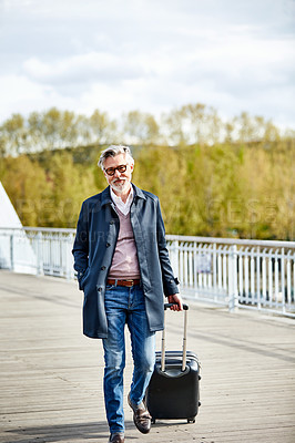 Buy stock photo Portrait of a mature man pulling a suitcase while walking over a bridge in the city