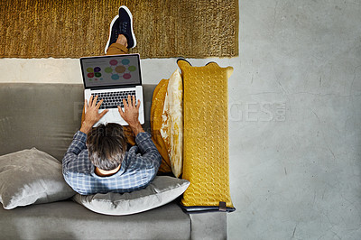Buy stock photo High angle shot of a young man working on a laptop while sitting on his sofa at home