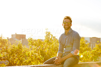 Buy stock photo Portrait of a smiling young man sitting on a rooftop at sunset with the city in the background