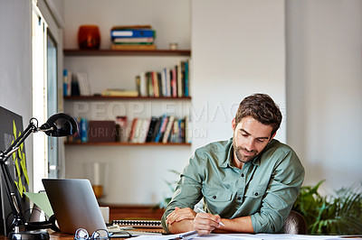Buy stock photo Shot of a young man sitting at a table at home working on a laptop