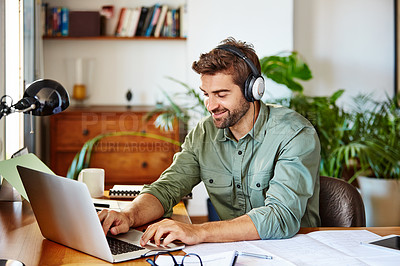 Buy stock photo Shot of a young man sitting at home wearing headphones and working on a laptop