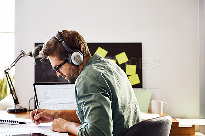 Buy stock photo Shot of a young man sitting at home wearing headphones and working on a laptop
