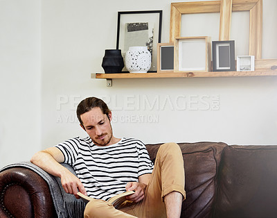 Buy stock photo Shot of a handsome young man reading a book while sitting on a sofa at home