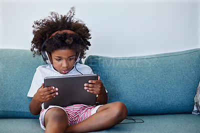 Buy stock photo Shot of a focused little boy browsing on a digital tablet and wearing headphones while relaxing on the couch at home