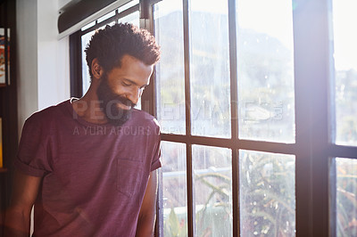 Buy stock photo Shot of a cheerful young man standing next to a window while looking down at the floor inside at home during the day