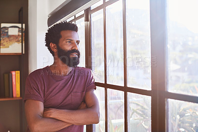 Buy stock photo Shot of a confident young man standing with his arms folded while looking through a window inside at home during the day