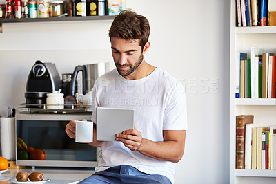 Buy stock photo Shot of a handsome young man using a digital tablet and having coffee in the morning at home