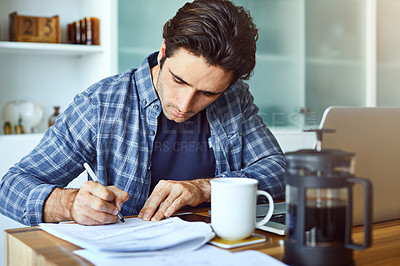 Buy stock photo Shot of a focused young man writing on a document while being seated behind his laptop at home