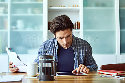 Buy stock photo Shot of a focused young man working on his digital tablet while being seated next to a table at home