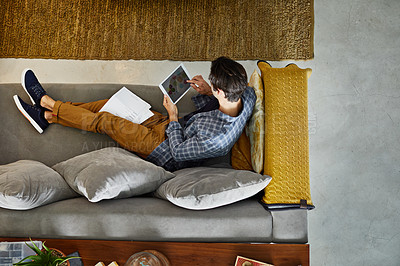 Buy stock photo High angle shot of a focused young unrecognizable man working on a digital tablet while relaxing on a couch at home