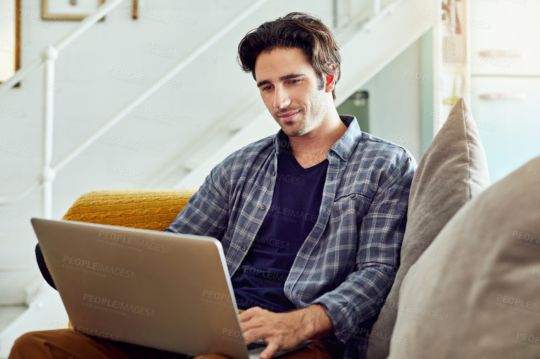 Buy stock photo Shot of a focused young man working on a laptop while being seated on a couch at home