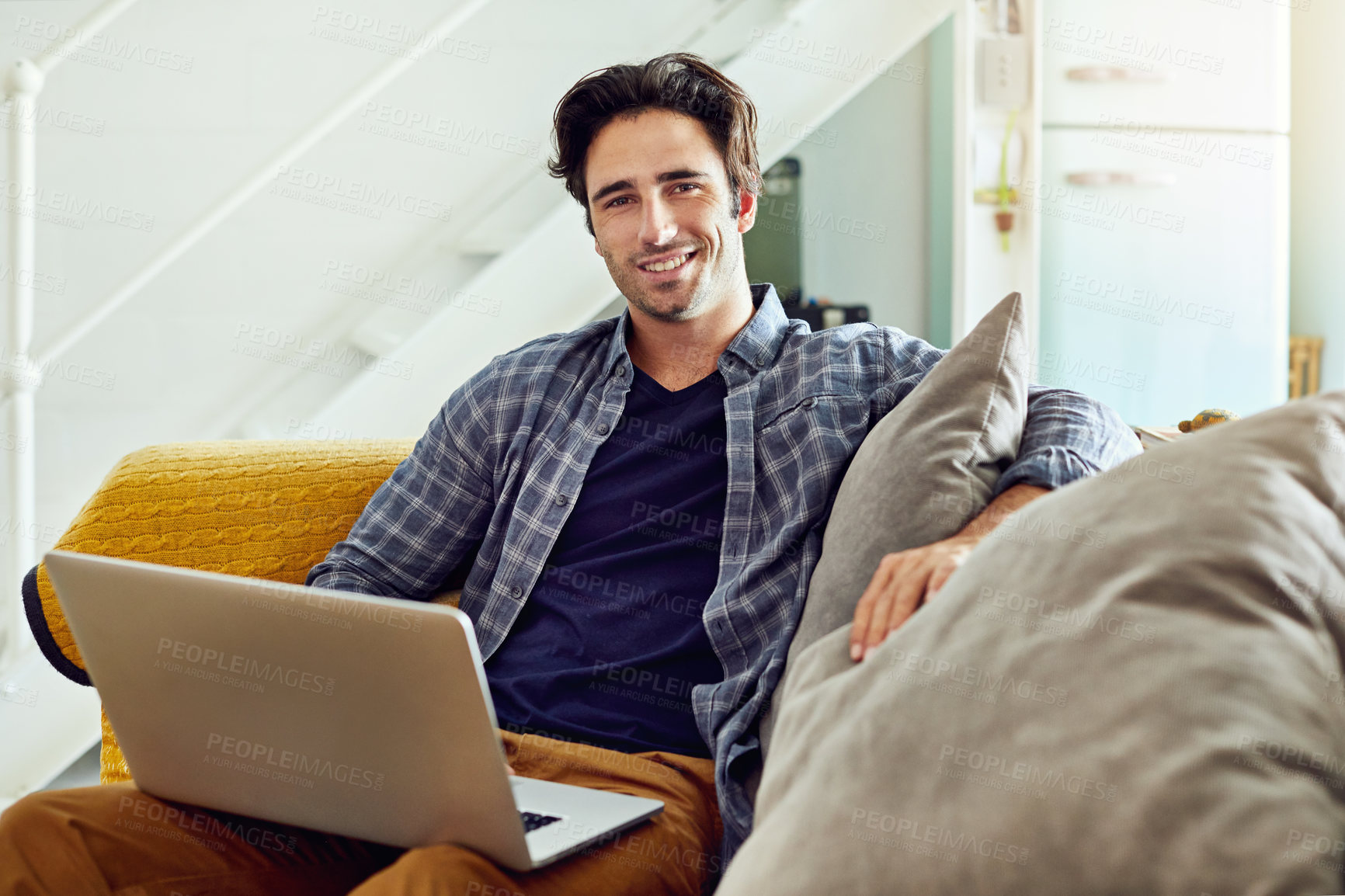 Buy stock photo Portrait of a cheerful young man working on a laptop while being seated on a couch at home