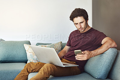 Buy stock photo Shot of a handsome young man using his laptop and mobile phone on the sofa at home
