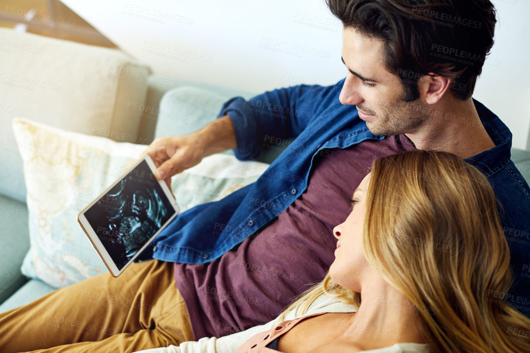 Buy stock photo Shot of a young man and his pregnant wife looking at a sonogram on a digital tablet on the sofa