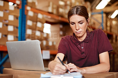 Buy stock photo Shot of a young woman writing notes while using a laptop in a warehouse