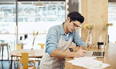 Buy stock photo Shot of a young cafe owner using his laptop and doing paperwork in his shop