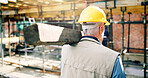 Back, man and lumber on construction site for renovation, scaffolding and building progress with wood. Mature male person, walking and plank for remodeling project, infrastructure upgrade with helmet