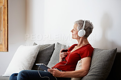 Buy stock photo Shot of a mature woman sitting on her sofa listening to music on a digital tablet and drinking wine