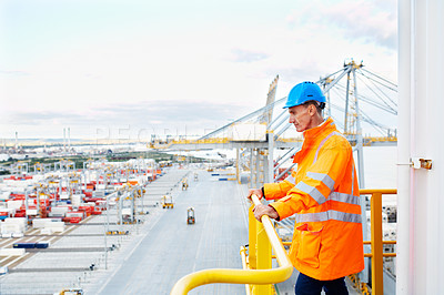 Buy stock photo Shot of a man in workwear standing on a walkway looking  out over at a large commercial dock
