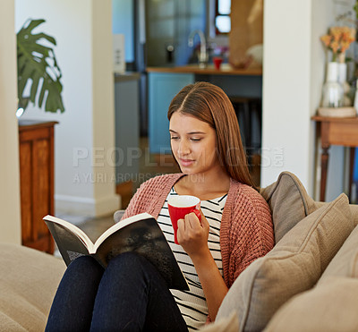 Buy stock photo Shot of a young woman sitting on her sofa reading a book and drinking a coffee
