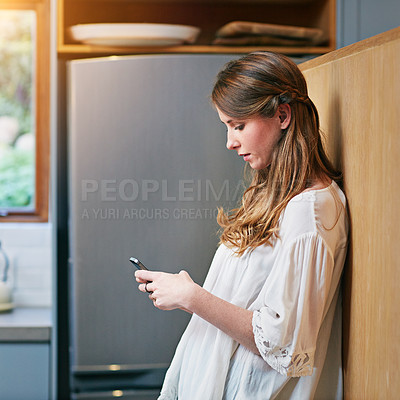 Buy stock photo Shot of a young woman using a cellphone in the kitchen