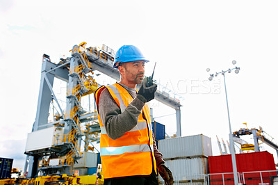 Buy stock photo Shot of a young man in workwear talking on a walkietalkie while standing on a large commercial dock
