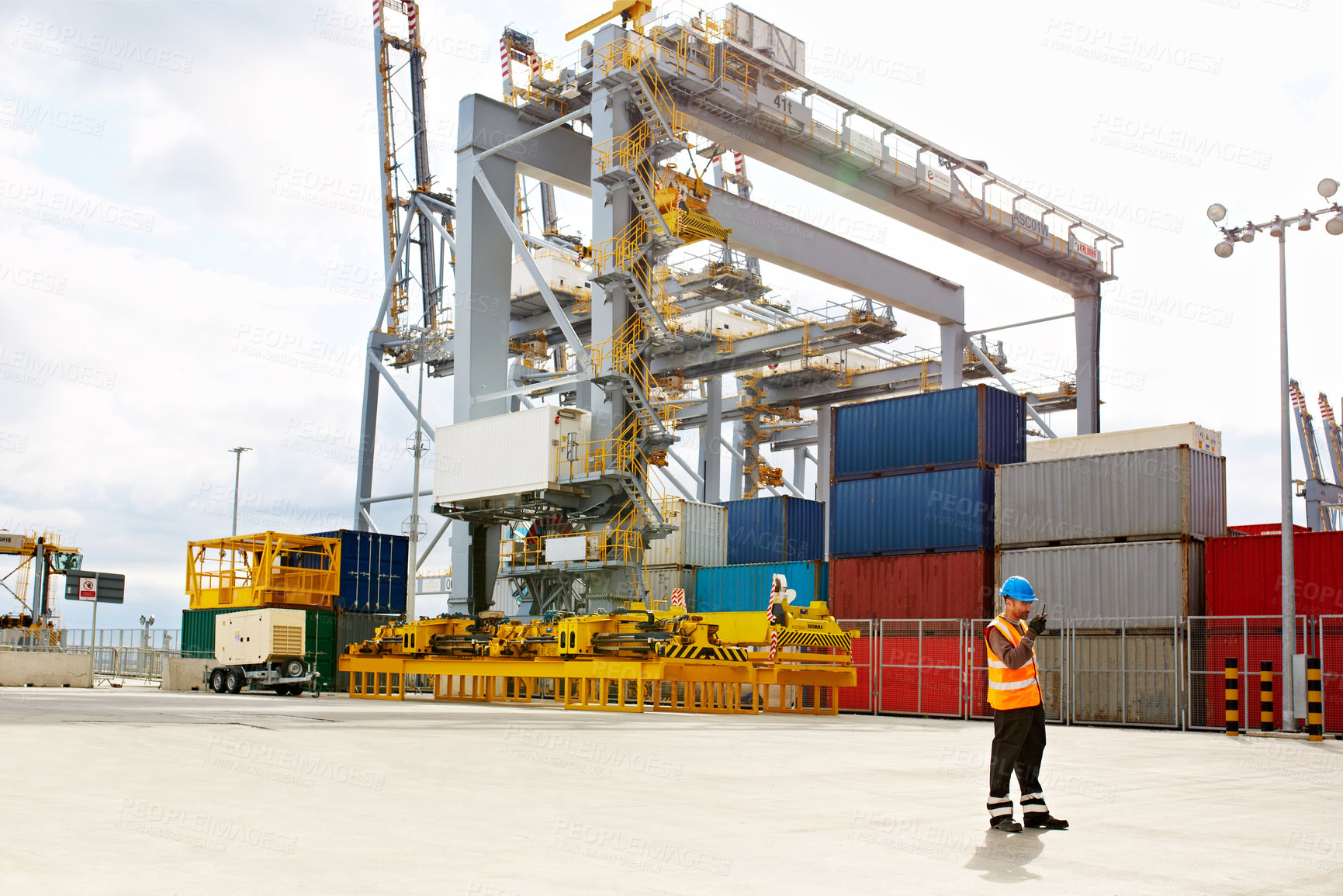 Buy stock photo Shot of a young man in workwear talking on a walkie talkie while standing on a large commercial dock