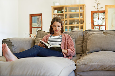 Buy stock photo Shot of a young woman sitting on her sofa reading a book