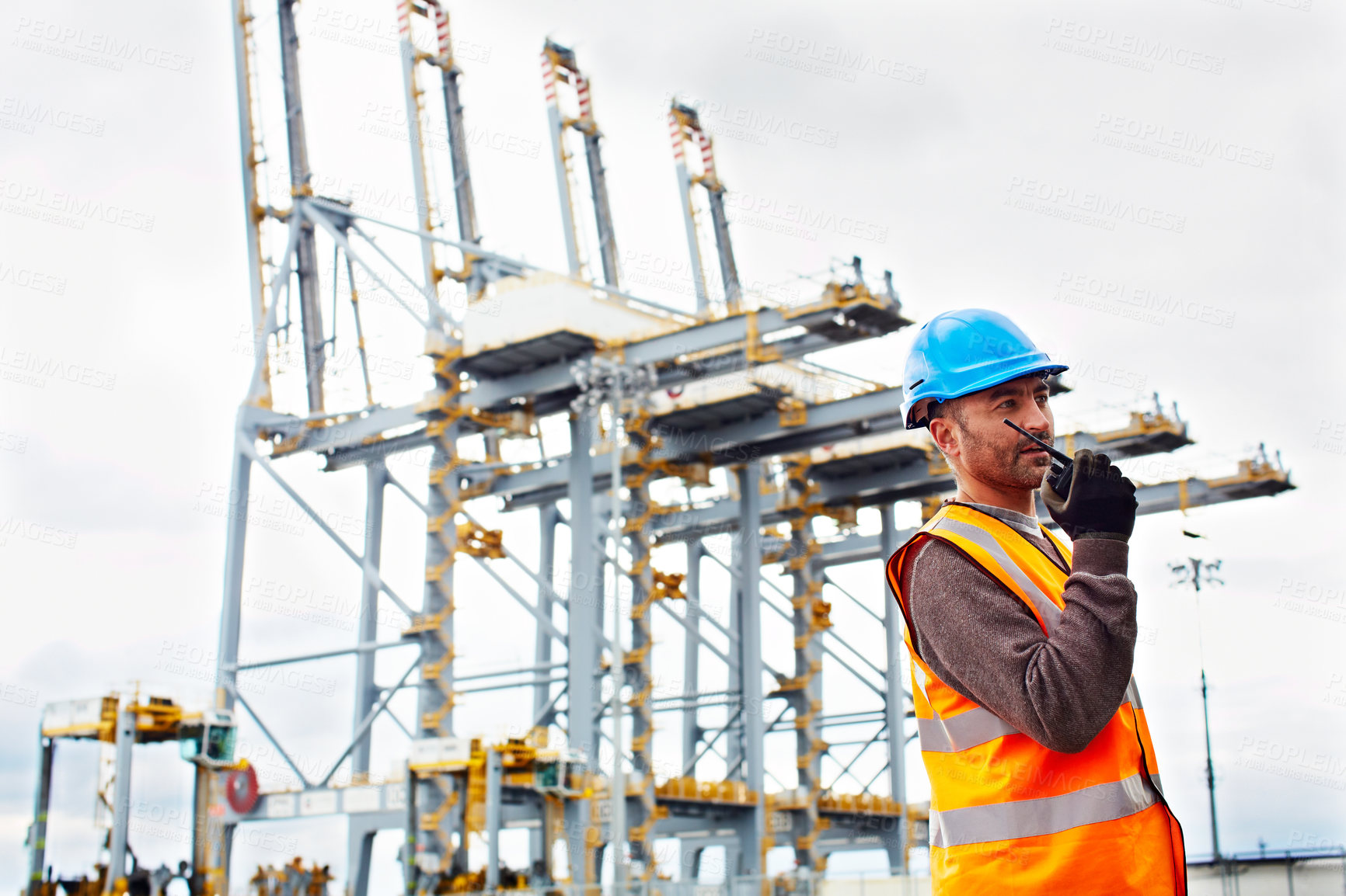 Buy stock photo Shot of a young man in workwear talking on a walkie talkie while standing on a large commercial dock