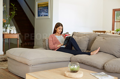 Buy stock photo Shot of a young woman sitting on her sofa reading a book and drinking a coffee