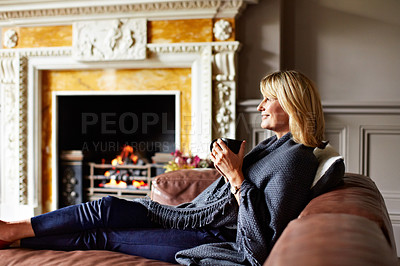 Buy stock photo Shot of a mature woman sitting on her sofa drinking tea while wrapped in a blanket