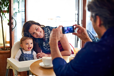 Buy stock photo Shot of a father taking a picture of his wife and baby girl while sitting in a cafe